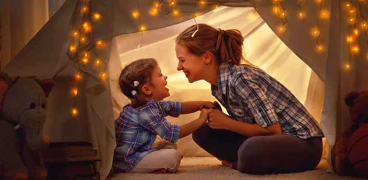 Dentro de una tienda de lona decorada con luces, una mujer y una niña pequeña se toman de las manos y sonríen.