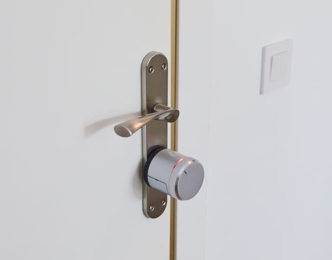 Primer plano de picaporte metálico moderno en puerta blanca, con cilindro electrónico plateado instalado debajo y un interruptor de luz visible al fondo.