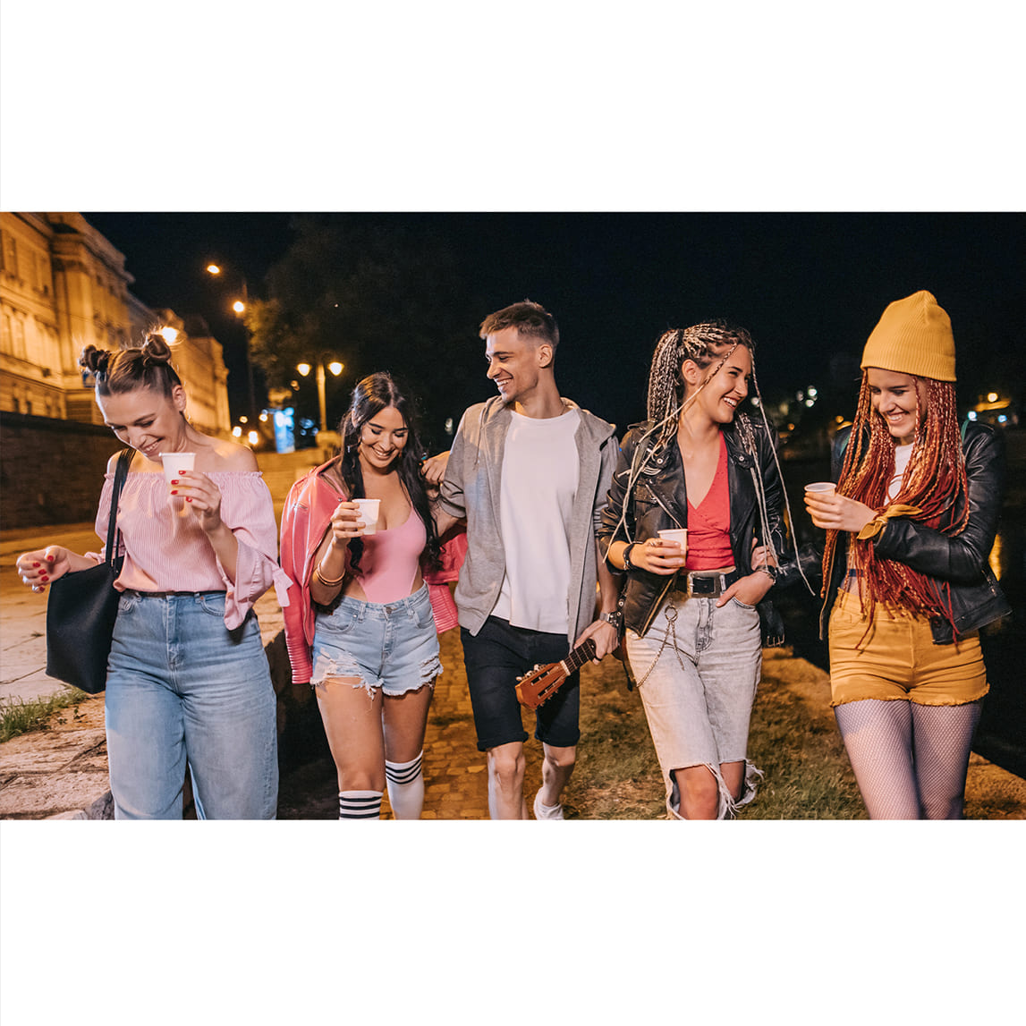 Grupo de cinco jóvenes caminando de noche por la calle mientras ríen y sostienen vasos.