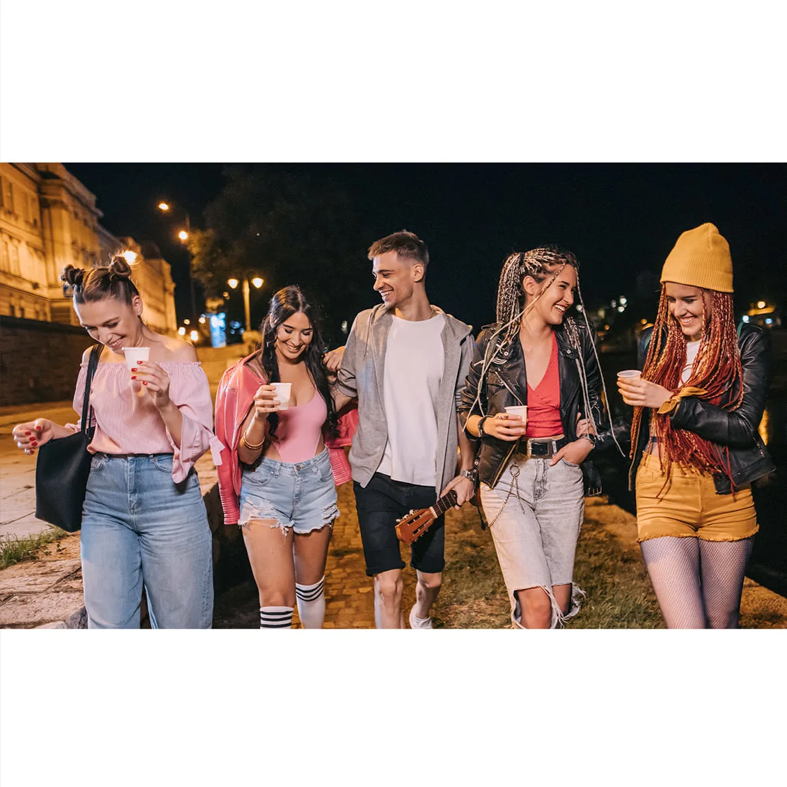 Grupo de cinco jóvenes caminando de noche por la calle mientras ríen y sostienen vasos.