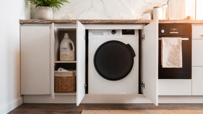 Lavadora integrable blanca instalada bajo encimera en cocina moderna, con puertas de armario abiertas, cesta de mimbre y detergente visibles junto a un horno