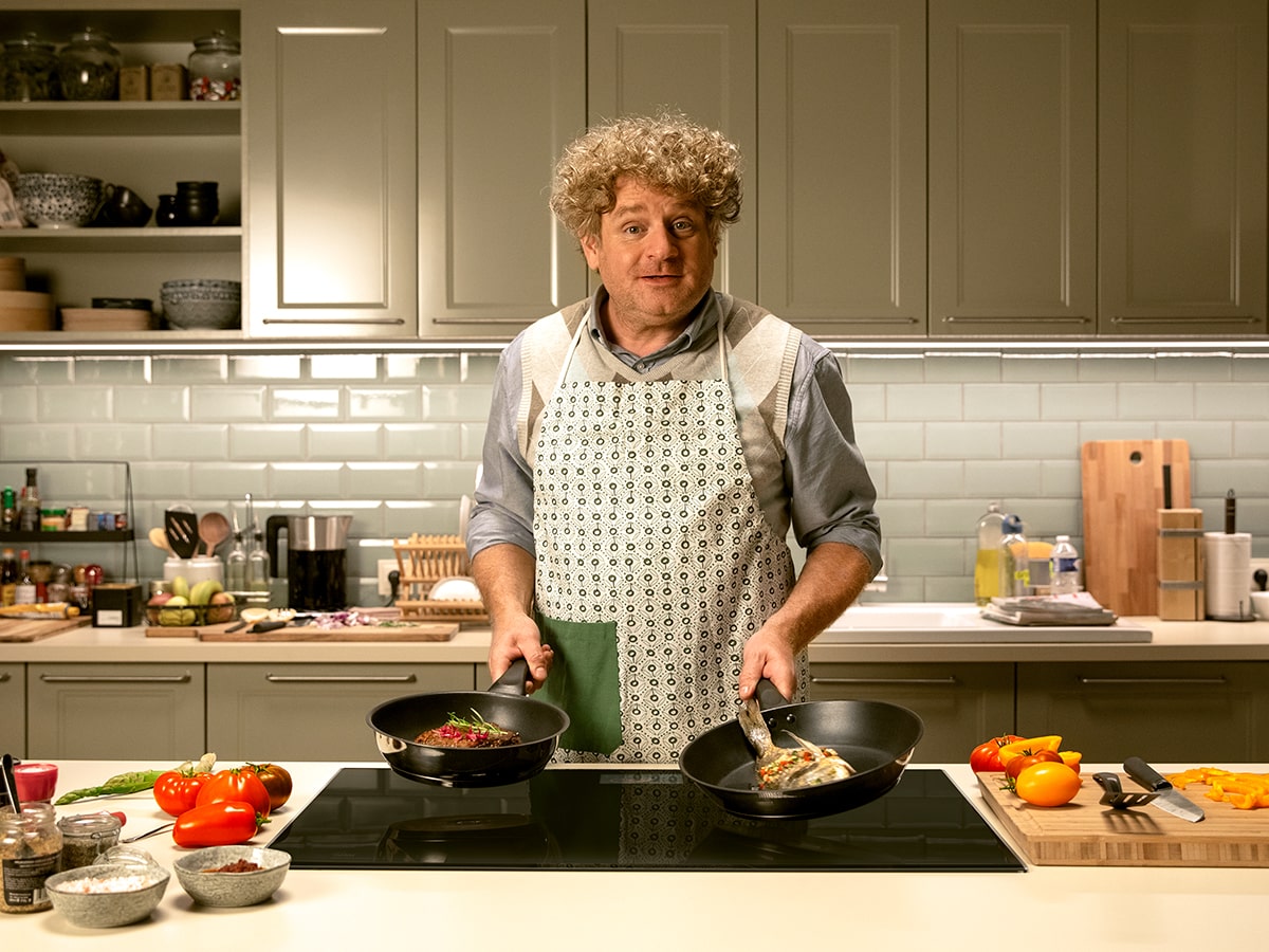 En una cocina, un hombre con delantal sostiene sartenes con comida sobre una placa. Hay armarios, utensilios y verduras.