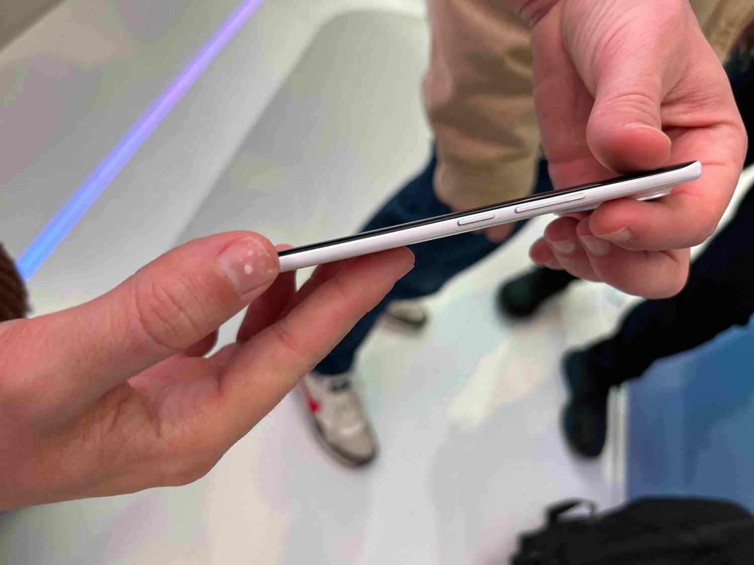 Mano sujetando un teléfono blanco con bordes negros, mostrando botones laterales. Fondo blanco con personas y zapatos visibles.