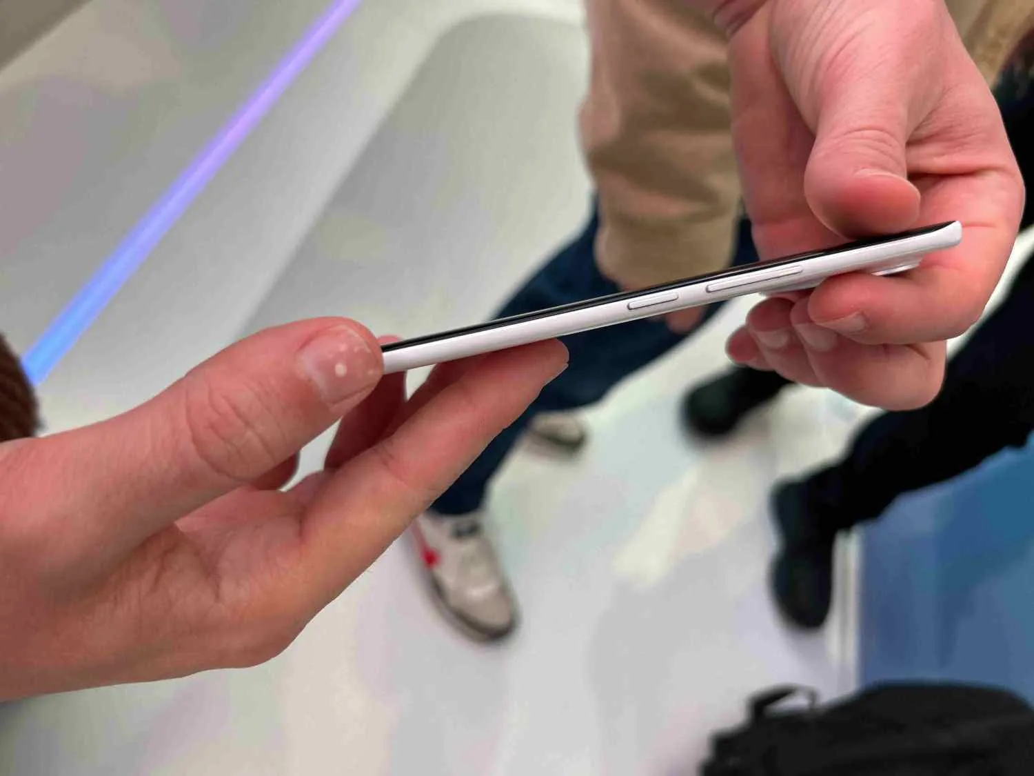 Mano sujetando un teléfono blanco con bordes negros, mostrando botones laterales. Fondo blanco con personas y zapatos visibles.