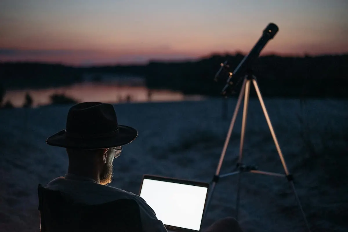Persona con portátil junto a un telescopio al atardecer.