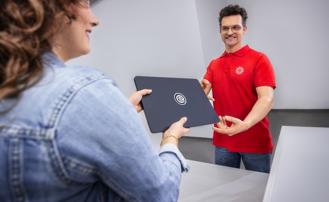 Una persona entrega un portátil negro con un logo a otra persona en una mesa, con un hombre de rojo al fondo.
