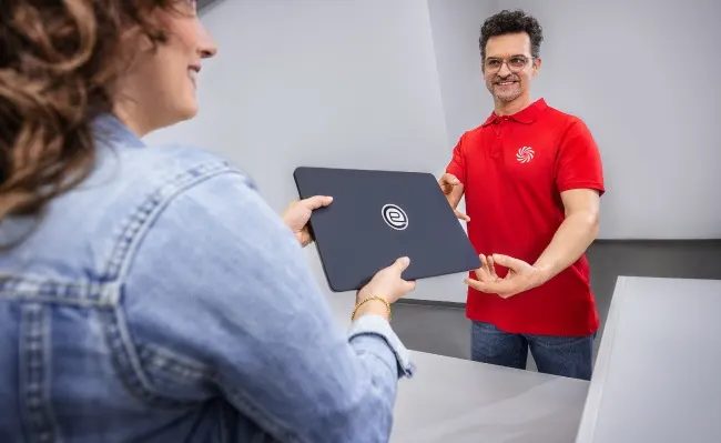 Una persona entrega un portátil negro con un logo a otra persona en una mesa, con un hombre de rojo al fondo.