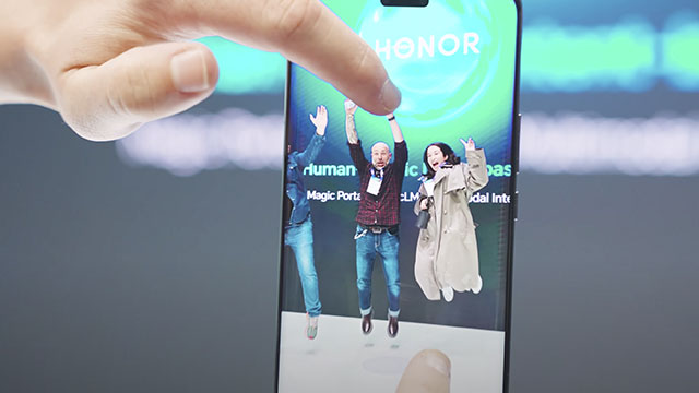 Un dedo toca la pantalla de un teléfono móvil. La pantalla muestra tres personas saltando sobre un fondo verde azulado.