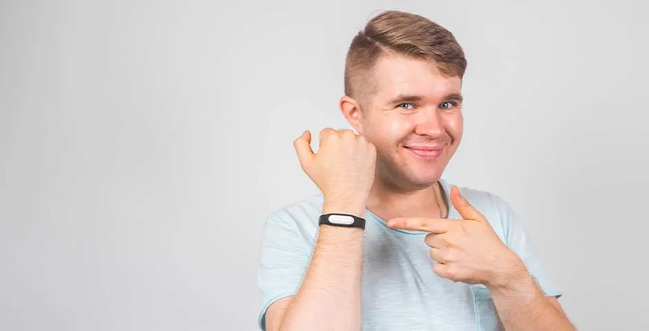 Un hombre sonriente señala una pulsera negra con pantalla en su muñeca izquierda, sobre un fondo gris claro.