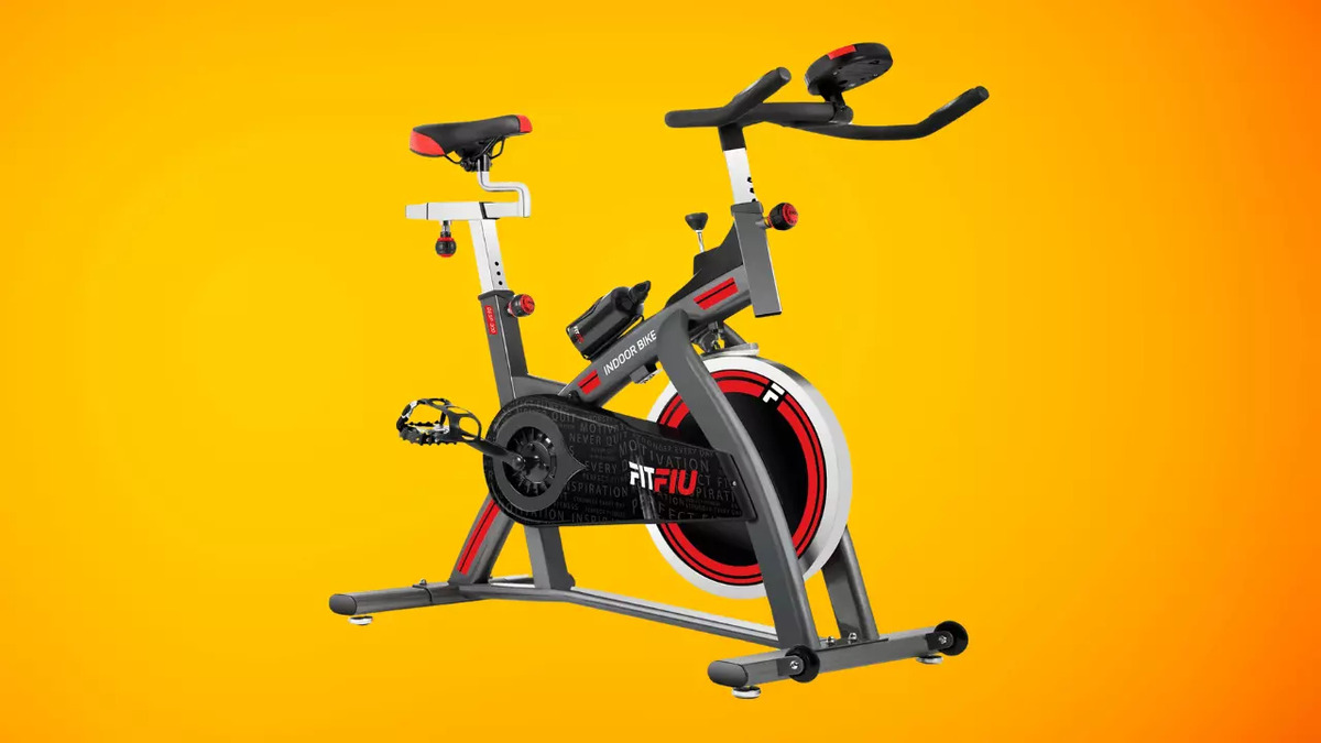 Bicicleta estática gris con detalles rojos, sillín rojo y manillar negro sobre un fondo amarillo. Tiene pedales negros.