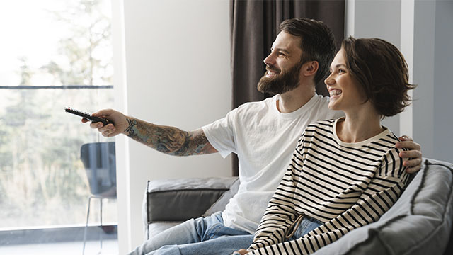 Una pareja sonriente sentada en un sofá gris, el hombre con un control remoto en la mano, mirando hacia una ventana grande.