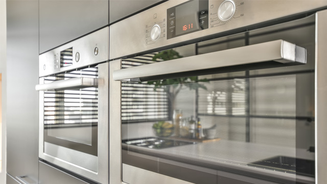 Dos hornos de acero inoxidable empotrados con panel de control y reflejo de cocina interior con persianas.