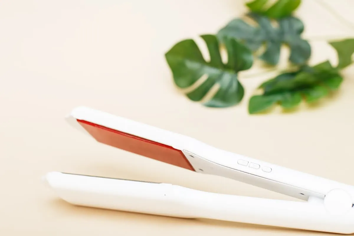 Plancha de pelo blanca sobre fondo beige con hojas verdes decorativas al fondo.