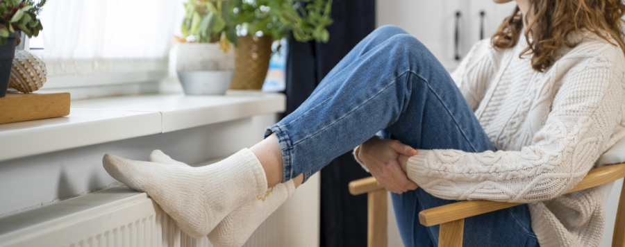Una persona sentada en una silla de madera, con jeans azules y calcetines blancos, apoyando los pies en un radiador blanco. En el fondo, plantas en macetas cerca de una ventana.
