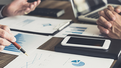 En una mesa de madera hay documentos con gráficos azules, un portátil, un móvil blanco y manos con un bolígrafo.