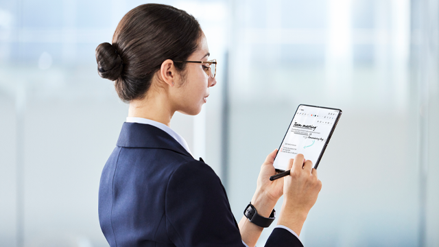 Mujer con gafas y traje azul, sujeta una tableta con un lápiz óptico en un entorno de oficina con fondo blanco.