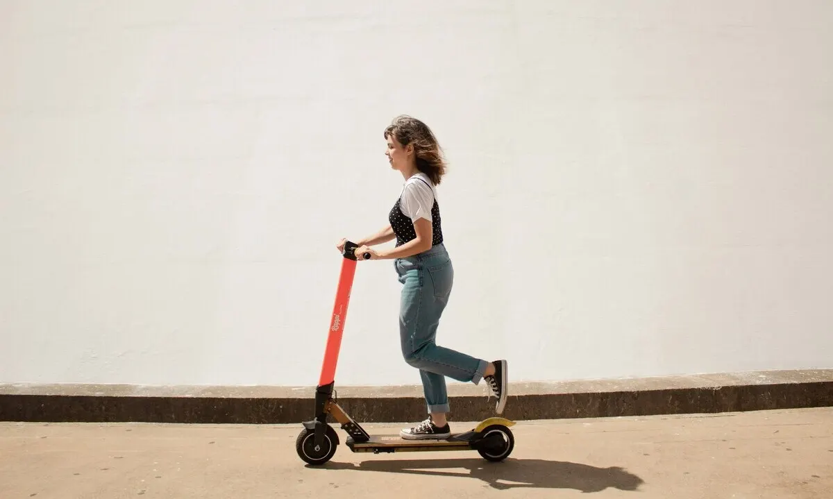 Mujer joven montando un patinete eléctrico rojo junto a una pared blanca.