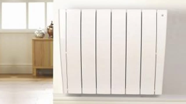 Un radiador blanco de pared con paneles verticales. Al fondo, un mueble de madera junto a una ventana rectangular.