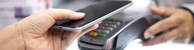 Mano sosteniendo un teléfono negro sobre un terminal de pago gris con botones verdes y amarillos, en un entorno de comercio.