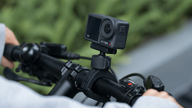 Cámara negra Action 4 montada en el manillar negro de una bicicleta, con fondo verde difuminado.