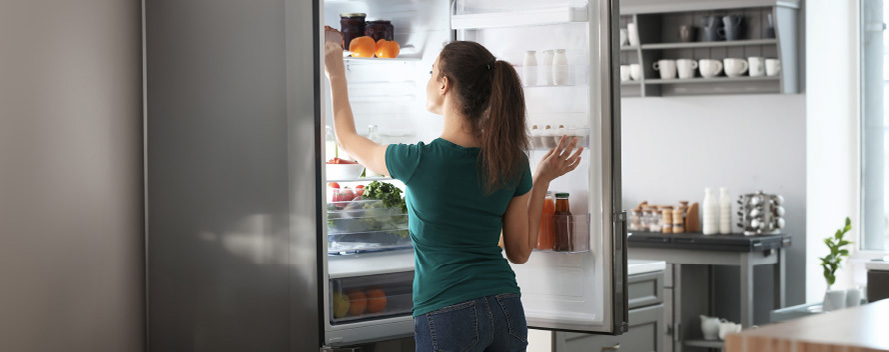 Una mujer abre un refrigerador plateado lleno de comida, incluyendo fruta y vegetales. Al fondo, se ve una estantería y una mesa.