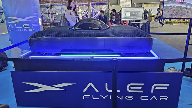 Coche volador "ALEF" sobre plataforma azul con logo. Personas y pantallas al fondo. Iluminación azulada en el entorno.