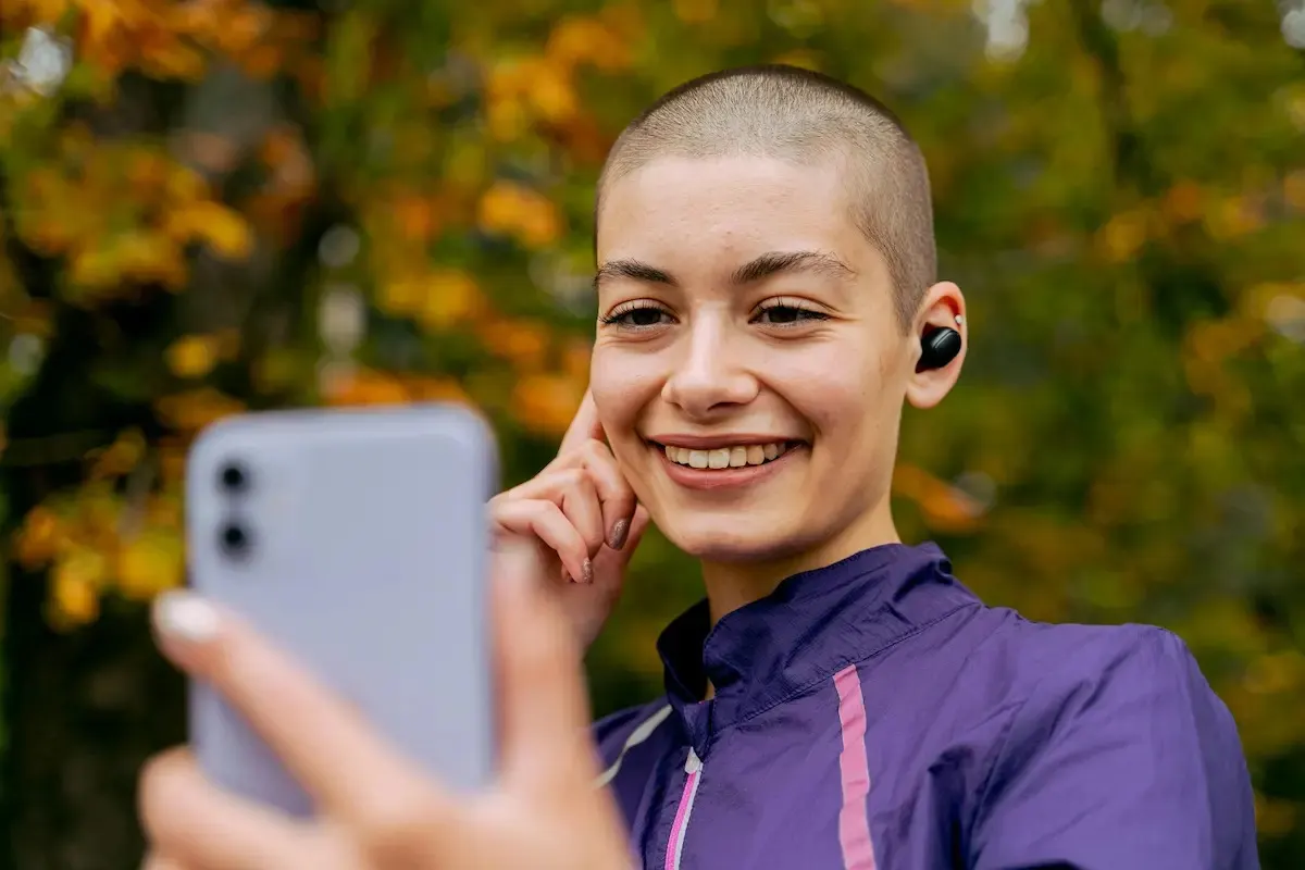 Una persona sonríe y sostiene un teléfono. Lleva audífonos y una chaqueta deportiva morada. El fondo muestra árboles.