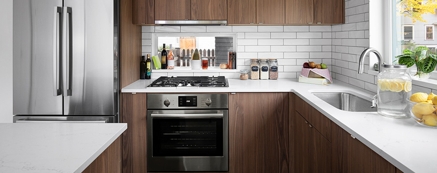 Cocina con nevera de acero, armarios de madera, encimera blanca con fregadero y horno de acero. Hay una ventana con plantas y limones.