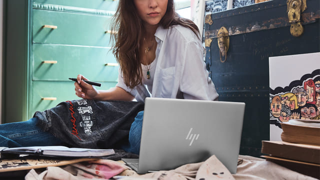 Mujer sentada en el suelo con portátil, cuaderno y bolígrafo. Al fondo, cómoda turquesa y baúl oscuro con detalles dorados.