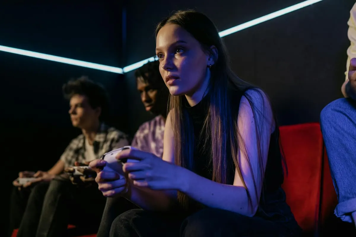 Imagen de una chica joven jugando con una PS5, sujetando un mando de color blanco. A su alrededor 3 personas que también están jugando.