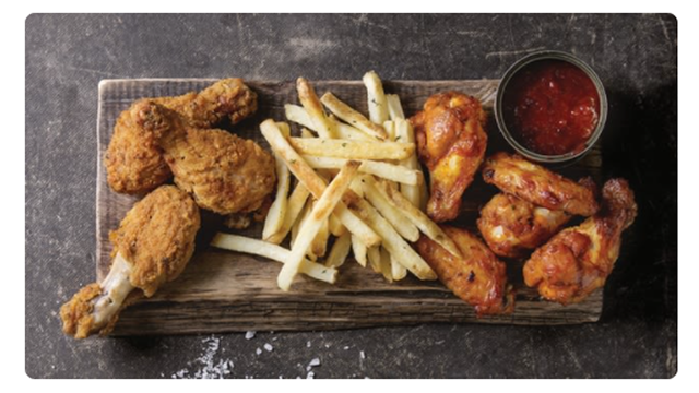 Sobre tabla de madera, hay pollo frito, papas fritas, salsa roja en un tazón. El fondo es gris oscuro.