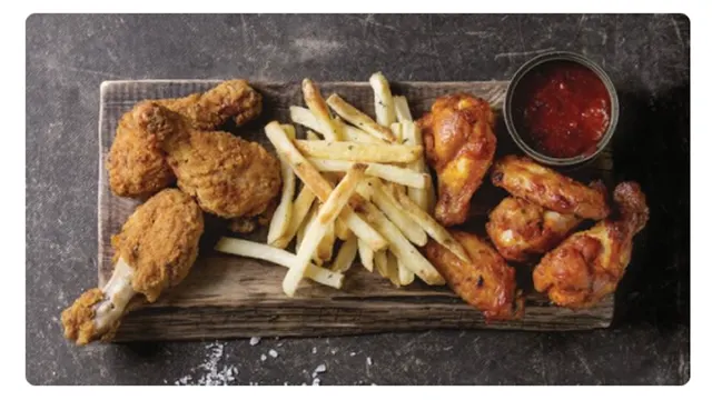 Sobre tabla de madera, hay pollo frito, papas fritas, salsa roja en un tazón. El fondo es gris oscuro.