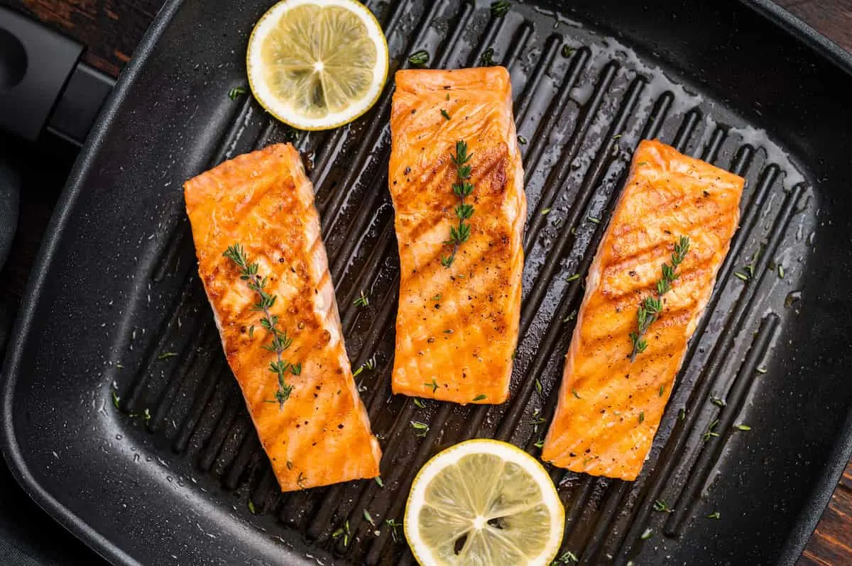 Tres filetes de salmón a la plancha con rodajas de limón y hierbas en una sartén negra acanalada.