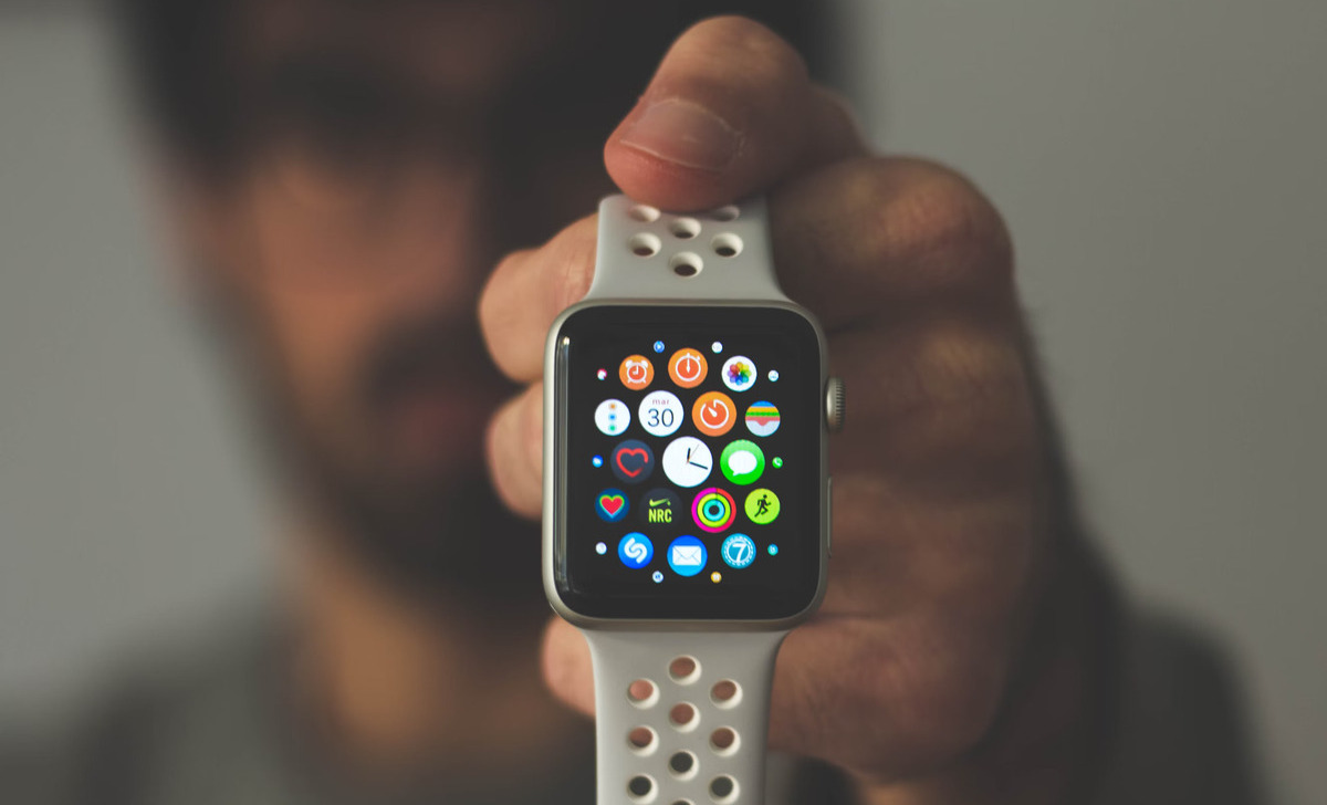 Mano sosteniendo un reloj inteligente con esfera cuadrada y correa blanca con orificios que muestra varios iconos de aplicaciones.