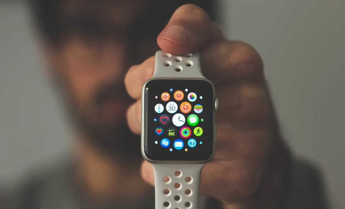 Mano sosteniendo un reloj inteligente con esfera cuadrada y correa blanca con orificios que muestra varios iconos de aplicaciones.