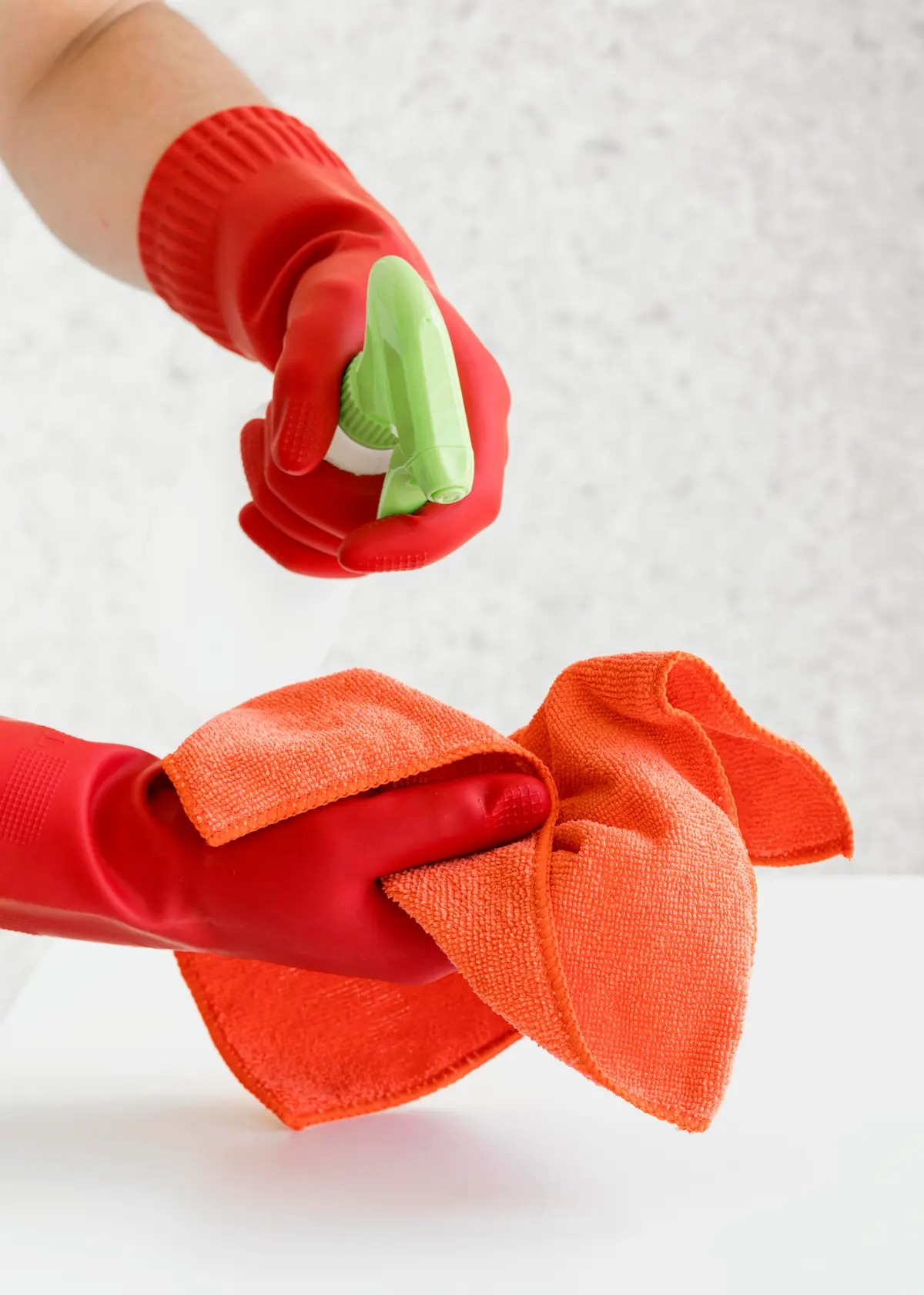 Manos enguantadas en rojo sostienen un atomizador verde y un paño de limpieza naranja sobre una superficie blanca.