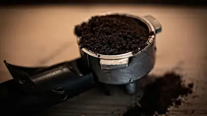 Un portafiltro de café metálico lleno de café molido sobre una superficie de madera. Parte del café está esparcido sobre la mesa.