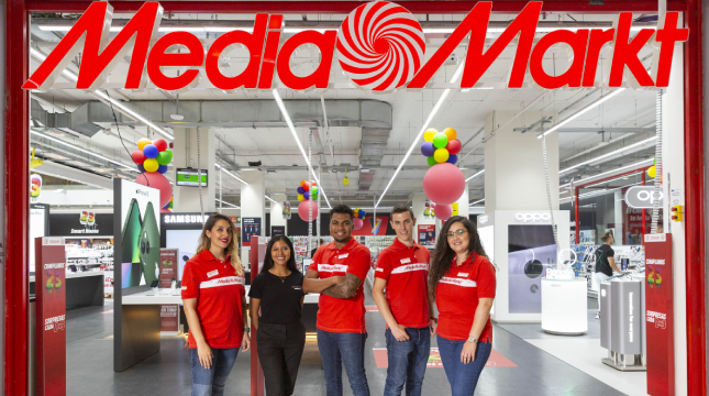 Un grupo de empleados delante de la tienda Media Markt. Globos en la tienda, luces en el techo y estantes con productos.