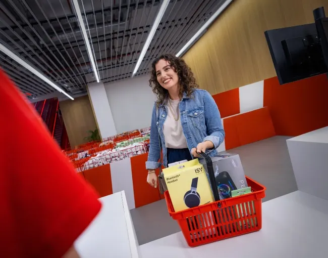 Una mujer sonríe mientras sostiene una cesta roja con una caja amarilla de auriculares en una tienda.