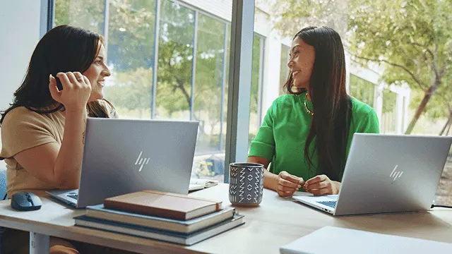 Dos mujeres sentadas en una mesa con computadoras portátiles, libros y una taza. Una lleva una camisa beige, la otra verde.