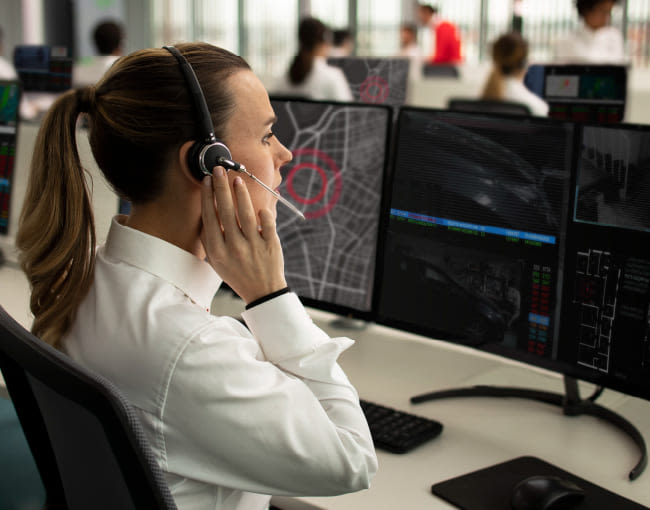 Operadora de central receptora con auriculares atiende llamada frente a varios monitores, mostrando mapas urbanos y datos de seguimiento en una sala moderna.

