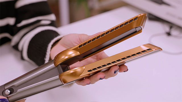 Una persona sostiene una plancha de pelo dorada y gris sobre una superficie blanca. La persona lleva un suéter a rayas.