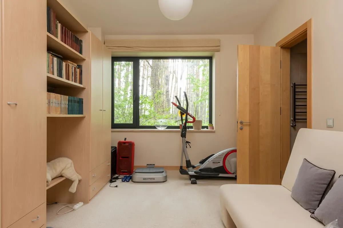 Habitación con sofá, estantería y máquina elíptica junto a la ventana.