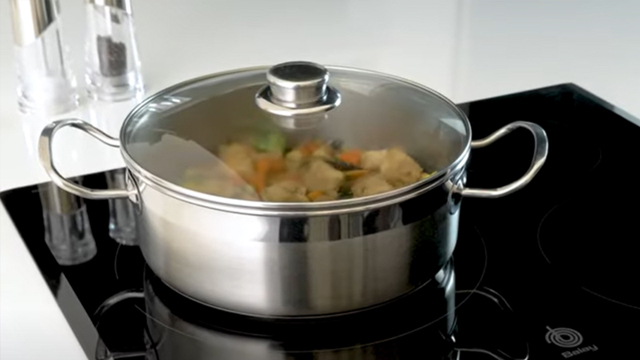 Recipiente de acero con tapa de cristal con alimentos cocinando hortalizas sobre una de las zonas de cocción.