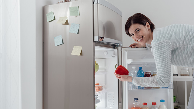 Una persona sonriente toma un pimiento rojo del refrigerador de acero inoxidable con notas adhesivas.