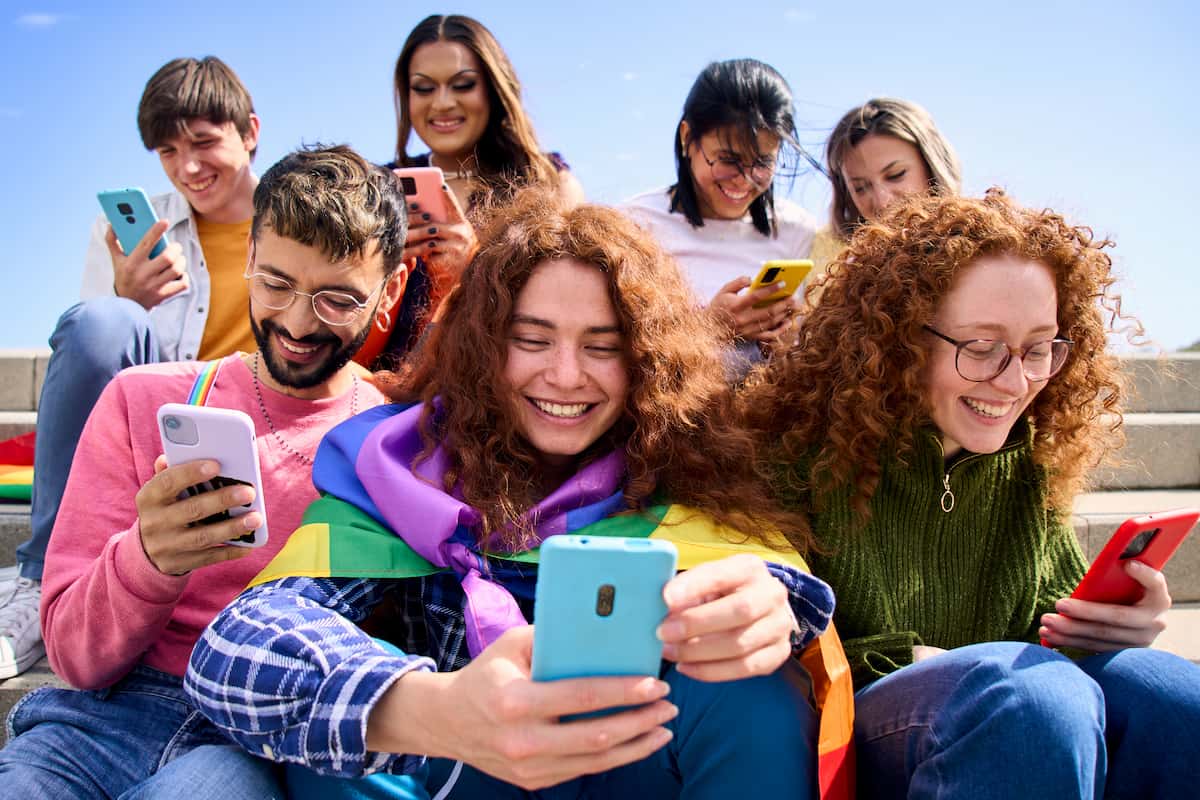 Grupo de jóvenes sonriendo mientras usan sus smartphones al aire libre, uno con bandera del arcoíris.