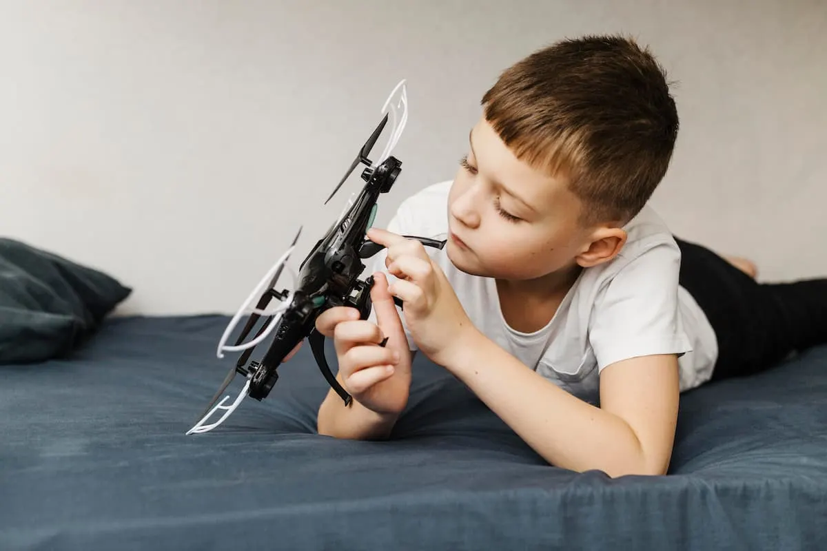 Niño tumbado en la cama examinando un dron entre sus manos.