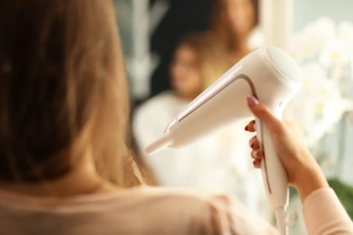 Persona de espaldas sosteniendo un secador de pelo blanco frente a un espejo, con el reflejo desenfocado y un fondo cálido.