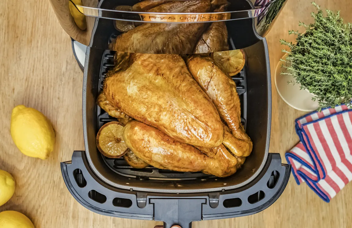 Vista superior de un pollo asado con rodajas de limón en una freidora de aire, sobre una mesa de madera con hierbas y un trapo.