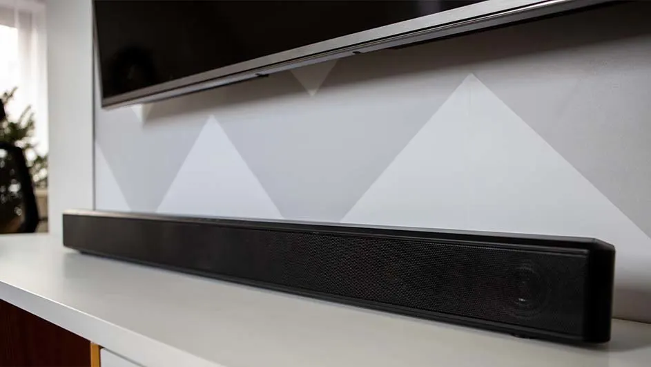 Televisor sobre pared con diseño de triángulos grises y blancos. Barra de sonido negra alargada sobre un mueble blanco.
